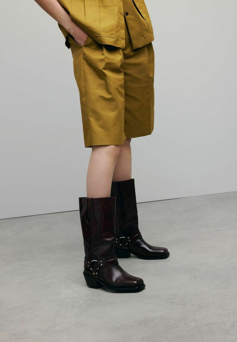 Bottes en cuir marron hautes jusqu'au genou avec des boucles, associées à un short de couleur moutarde. Le tissu est lisse avec une coupe décontractée.
