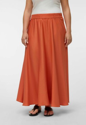 Femme portant une longue jupe fluide orange rouille avec une ceinture élastique et des sandales noires à bouts ouverts, debout devant un fond blanc.