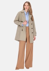Gabardina beige con diseño de doble botonadura, solapas grandes y mangas con puños. Llevada sobre una camisa azul claro y pantalones sueltos marrones.