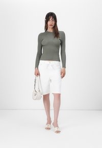 Top lungo a costine in olive, pantaloni corti bianchi fino al ginocchio, sandali beige con logo, mentre tiene una piccola borsa beige con hardware dorato.