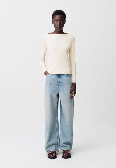 Donna che indossa un top a maniche lunghe color crema, jeans a palazzo blu chiaro, sandali neri con dettagli a catena e un grande anello d'oro sulla mano destra.
