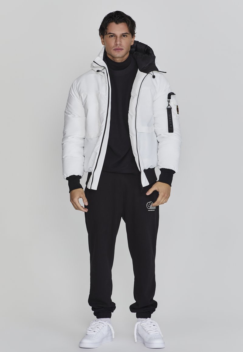 Hvid polstret jakke med hætte i sort, lynlås foran og sidelommer, parret med sorte joggingbukser og hvide sneakers. Enkel design, ingen mønstre.