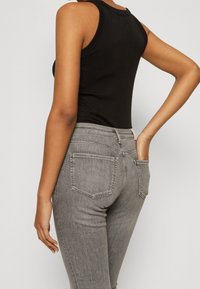 Persona vestida con una camiseta sin mangas negra y jeans de talle alto grises, con una mano en el bolsillo trasero, frente a un fondo liso.