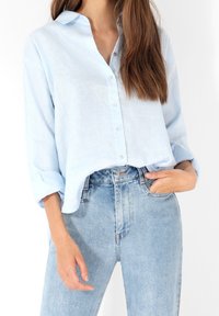 Chemise bleu clair à manches longues, boutonnée, fabriquée dans un tissu texturé, accompagnée d'un jean en denim clair taille haute. Détail de col simple visible.