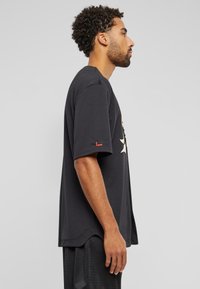 Tricou negru din bumbac cu mâneci scurte, având un imprimeu grafic mare pe față și un accent roșu pe mânecă. Vedere din lateral.