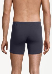 Man i mörkgråa figurnära boxerbriefs, sedd bakifrån med bar överkropp och armarna avslappnat vid sidorna.