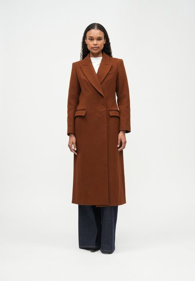 Woman wearing a long brown tailored coat, white top, wide-leg dark jeans, standing against a plain white background.