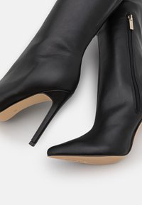 Botas de tacón alto de cuero negro con puntera puntiaguda y silueta ajustada. Cuentan con un cierre lateral de cremallera y una textura lisa y elegante.