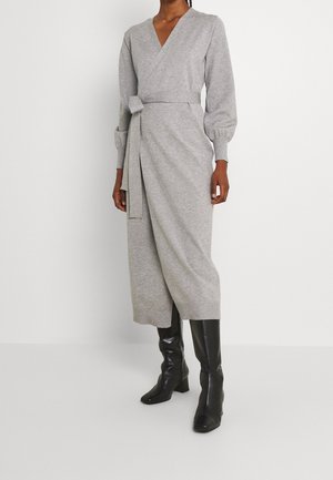 Kobieta ubrana w szarą sukienkę kopertową z długim rękawem i wiązaniem z boku, oraz czarne wysokie kozaki na obcasie, stoi na tle jednolitego białego tła.