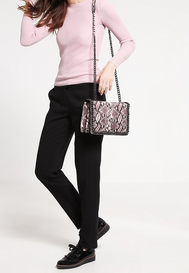 Donna che indossa un top a maniche lunghe a coste rosa, pantaloni neri, scarpe nere, mentre tiene una piccola borsa a tracolla in stampa pelle di serpente rosa con una tracolla nera.