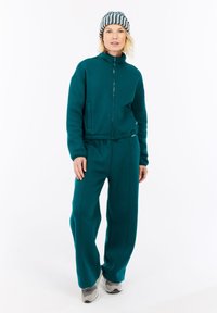 Una donna che indossa una giacca zip-up turchese e pantaloni a gamba larga, sneakers grigie e un berretto a righe, in piedi davanti a uno sfondo bianco.