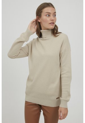 Pull à col roulé beige avec un motif texturé, des poignets et un ourlet côtelés. Porté avec un pantalon ajusté marron. Détail de marque minimal au niveau de l'ourlet.