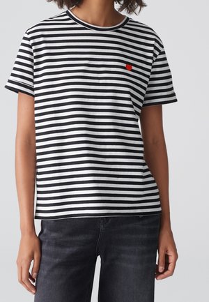 Mujer con una camiseta de rayas horizontales negras y blancas de manga corta con un pequeño corazón rojo en el pecho izquierdo, combinada con vaqueros negros.