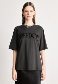 Μαύρο βαμβακερό t-shirt με κοντά μανίκια που διαθέτει μια μεγάλη γραφική παράσταση με την λέξη "ΕΛΕΥΘΕΡΙΑ" σε μαύρο χρώμα, φορεμένο με ασημένια αλυσίδα.