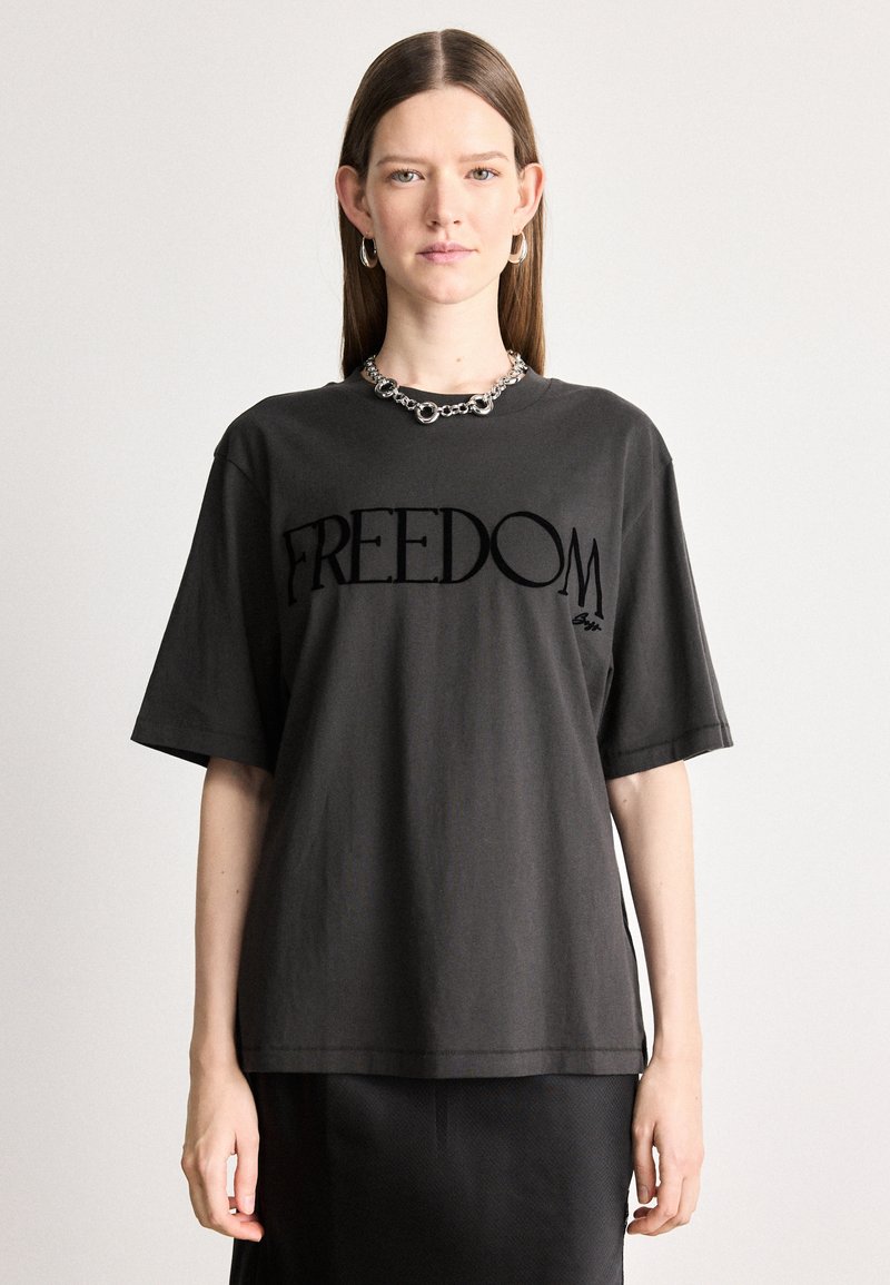 Μαύρο βαμβακερό t-shirt με κοντά μανίκια που διαθέτει μια μεγάλη γραφική παράσταση με την λέξη "ΕΛΕΥΘΕΡΙΑ" σε μαύρο χρώμα, φορεμένο με ασημένια αλυσίδα.