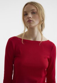 Rotes geripptes Langarmshirt mit breitem Rundhalsausschnitt, das eine taillierte Passform und eine weiche Textur aufweist. Einfaches Design ohne sichtbare Muster oder Akzente.