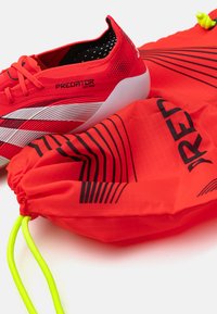 Red athletic shoes with white stripes and textured soles, alongside a matching red drawstring bag featuring black accents and a neon green cord.