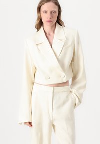 Blazer corto crema y pantalones a juego, hechos de una tela texturizada con patrones florales en relieve. Presenta solapas amplias y botones dobles.