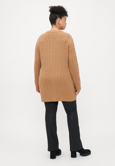 Long gilet en maille torsadée marron avec un ourlet côtelé. Porté par-dessus un pantalon noir, avec des manches longues et une coupe décontractée.