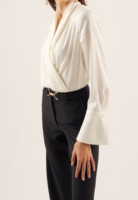 Weiße Wickelbluse mit langen, glockenförmigen Ärmeln, kombiniert mit hoch taillierten schwarzen Hosen mit einem Gürtel mit goldener runder Schnalle.