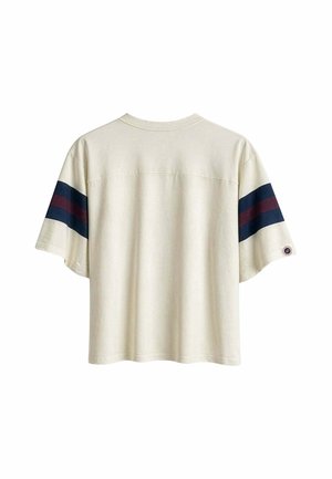 Camiseta beige de manga corta con rayas horizontales en azul marino y rojo oscuro en ambas mangas y una pequeña parche circular en la manga derecha.