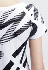 Bílá tričko s kombinací černých geometrických vzorů, včetně pruhů a teček, s kulatým výstřihem a krátkými rukávy.