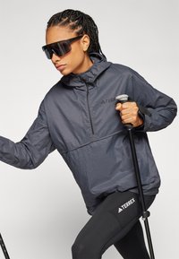 Jachetă ușoară, gri, cu fermoar la jumătate, având un model texturat și glugă, purtată cu colanți negri care au un logo. Utilizatorul ține bețe de trekking.