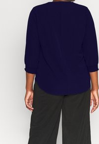 Blusa blu navy con maniche a tre quarti, presenta dettagli arricciati ai polsi, scollatura rotonda e un orlo curvo. Indossata con pantaloni larghi scuri.