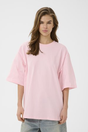 Woman with long brown hair wears oversized light pink T-shirt and light blue jeans, standing against plain white background.