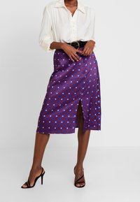 Une jupe mi-longue avec un motif à carreaux bleu et rouge, ornée d'accents floraux, dotée d'une fente à l'avant et d'une finition lisse et brillante.