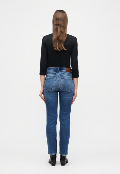 Femme debout de dos portant un haut noir, un jean taille haute bleu et des bottines noires, sur un fond blanc uni.