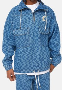 Veste en denim bleu avec un motif géométrique, deux poches poitrine, un devant à demi-zip, un ourlet réglable avec cordon de serrage, et un patch logo sur la poitrine.