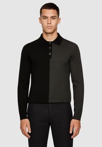 Uomo che indossa una polo a maniche lunghe bicolore nera e grigia scura con tre bottoni, abbinata a pantaloni neri, in piedi contro uno sfondo grigio.