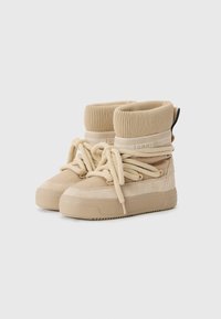 Beige ankle boots with a ribbed knit cuff, suede and synthetic material, round toe, flat sole, and pronounced laces. Featuring brand logo detail.