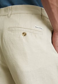 Pantalons en lin beige avec une poche arrière, dotés d'un bouton marron et d'une étiquette blanche affichant "Marc O'Polo." Texture lisse, design ajusté.