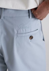 Light blue pants made of fabric with a smooth texture, featuring an embroidered "Prohibited" detail and a single brown button on the back pocket.