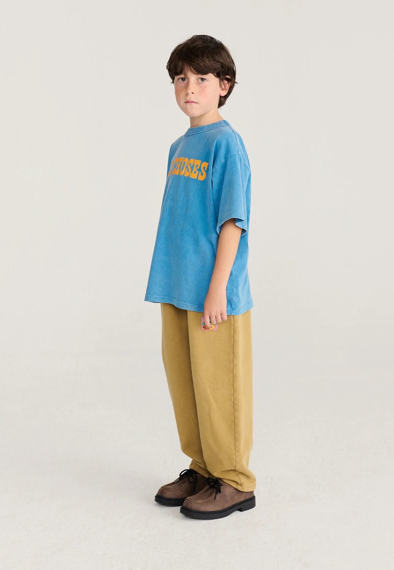 Garçon aux cheveux courts et foncés portant un t-shirt bleu oversize avec un texte jaune, un pantalon beige large et des chaussures marron à lacets, debout sur un fond clair.