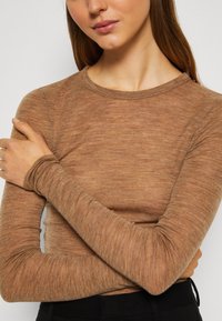 Haut marron à manches longues en tissu doux et texturé. Présente un col rond et des manches ajustées, montré en gros plan avec les bras croisés.