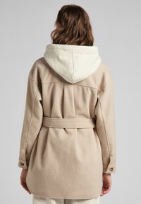 Mujer con cabello castaño ondulado que lleva un abrigo beige con cinturón y capucha crema, vista desde atrás contra un fondo gris liso.