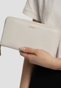 Cartera de cuero gris claro con textura de grano, cierre de cremallera y logo dorado "LAZAROTTI" en la parte frontal, sujetada en una mano.