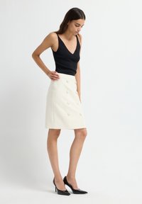 Zwarte V-hals tanktop en crèmekleurige A-lijn rok met opvallende knoopdetails. Accessoires zijn zwarte puntige hakken. Soepele texturen.