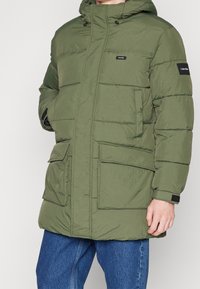 Mand iført en olivengrøn polstret vinterjakke med hætte, forsyninger lommer, og Calvin Klein-logoer på brystet og ærmet, parret med blå jeans.