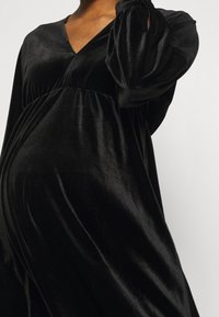 Černé sametové těhotenské šaty s výstřihem do V, volným střihem a balónovými rukávy, s nařasenými detaily v pase pro pohodlí.