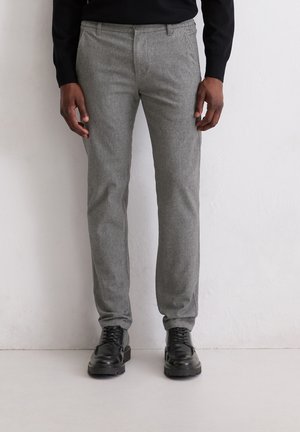 Mann trägt schlanke graue Hose, schwarze Lederschnürschuhe und einen schwarzen Pullover, steht vor einer weißen Wand.