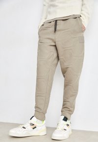 Calças de moletão beges com um padrão hexagonal texturizado, cintura elástica e bolsos laterais, combinadas com ténis brancos com detalhes em neon.