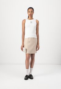 Weißes ärmelloses Oberteil mit Logodetail, kombiniert mit einem beigen, strukturierten Rock mit Metallelementen und schwarzen Loafers mit weißen Socken.