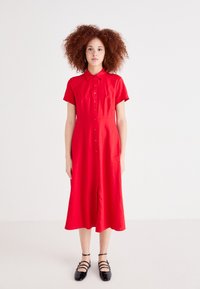 Κόκκινο φόρεμα με κουμπιά και κοντά μανίκια, μέχρι το γόνατο, φτιαγμένο από ελαφρύ ύφασμα. Μαύρες σαμπό με λουριά αναδεικνύουν την εμφάνιση.