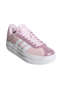 Zapatilla rosa con detalles de ante y acabados brillantes, que presenta rayas blancas, una punta texturizada y una suela blanca gruesa. Los cordones son de color rosa claro.