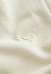 Tissu couleur crème présentant un tissage texturé avec un logo brodé subtil dans une teinte plus claire. Texture lisse et douce avec une légère brillance.