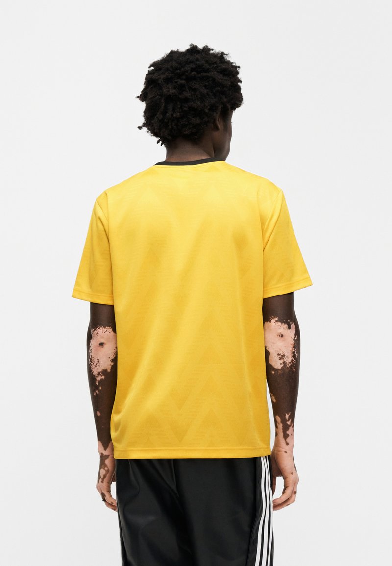 T-shirt jaune à manches courtes avec un motif texturé en zigzag et un col noir, associé à un short de sport noir.
